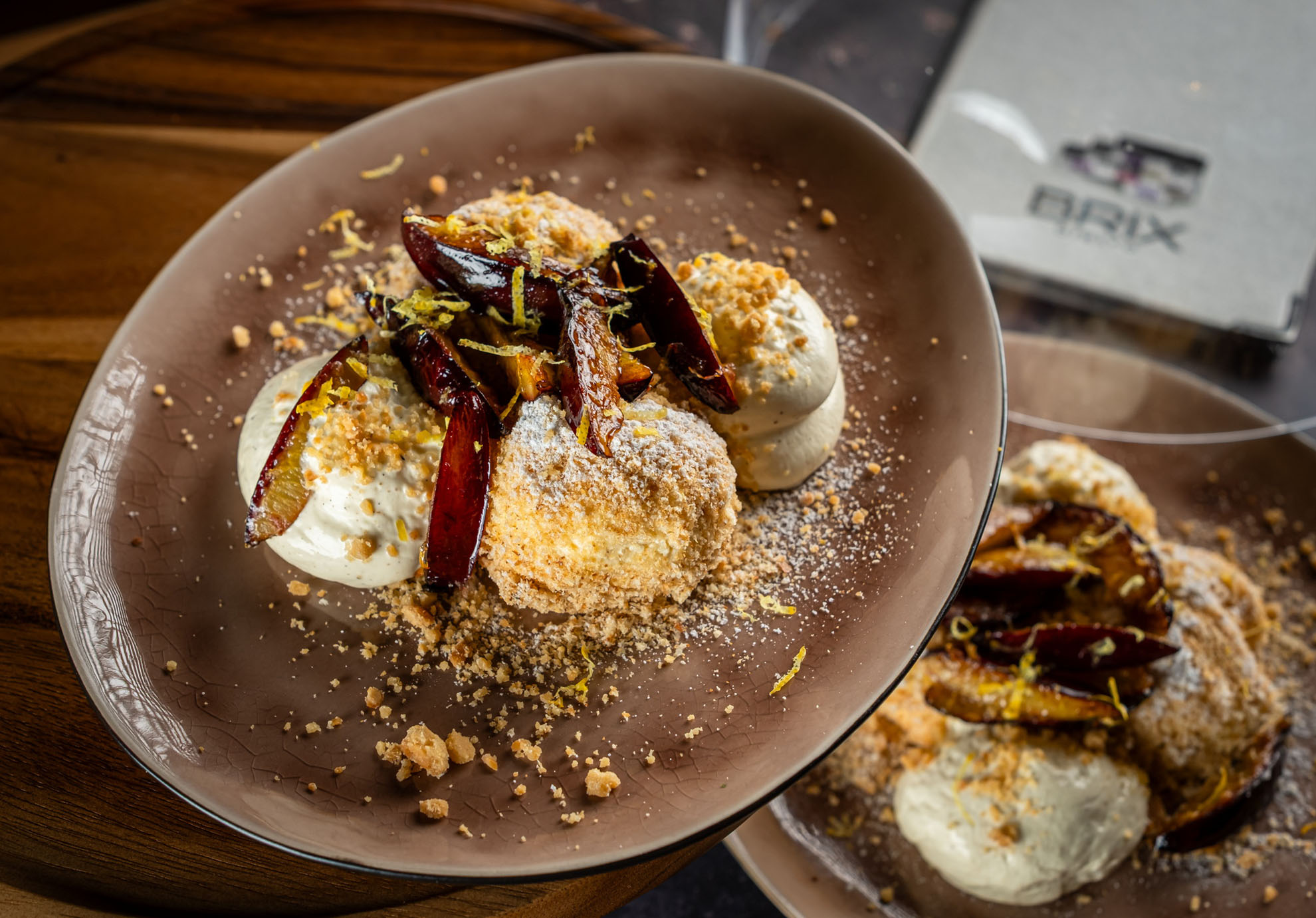 Egy rusztikus desszert barna tányéron, krémes túrógombóc, karamellizált fügével, morzsás szórás és citromhéjjal, egy faasztalon.