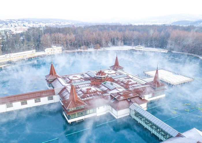 Luftaufnahme des Hévízer Sees im Winter mit dampfendem Thermalwasser, dem historischen Seebadgebäude und der verschneiten Landschaft.